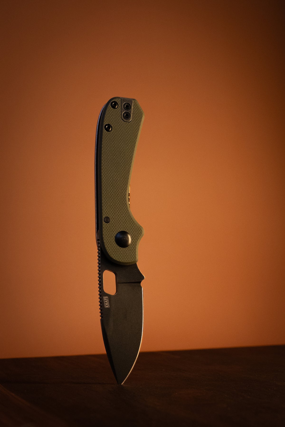 A folding pocket knife with a textured olive green handle standing vertically on a dark wooden surface against a warm orange-brown gradient background.