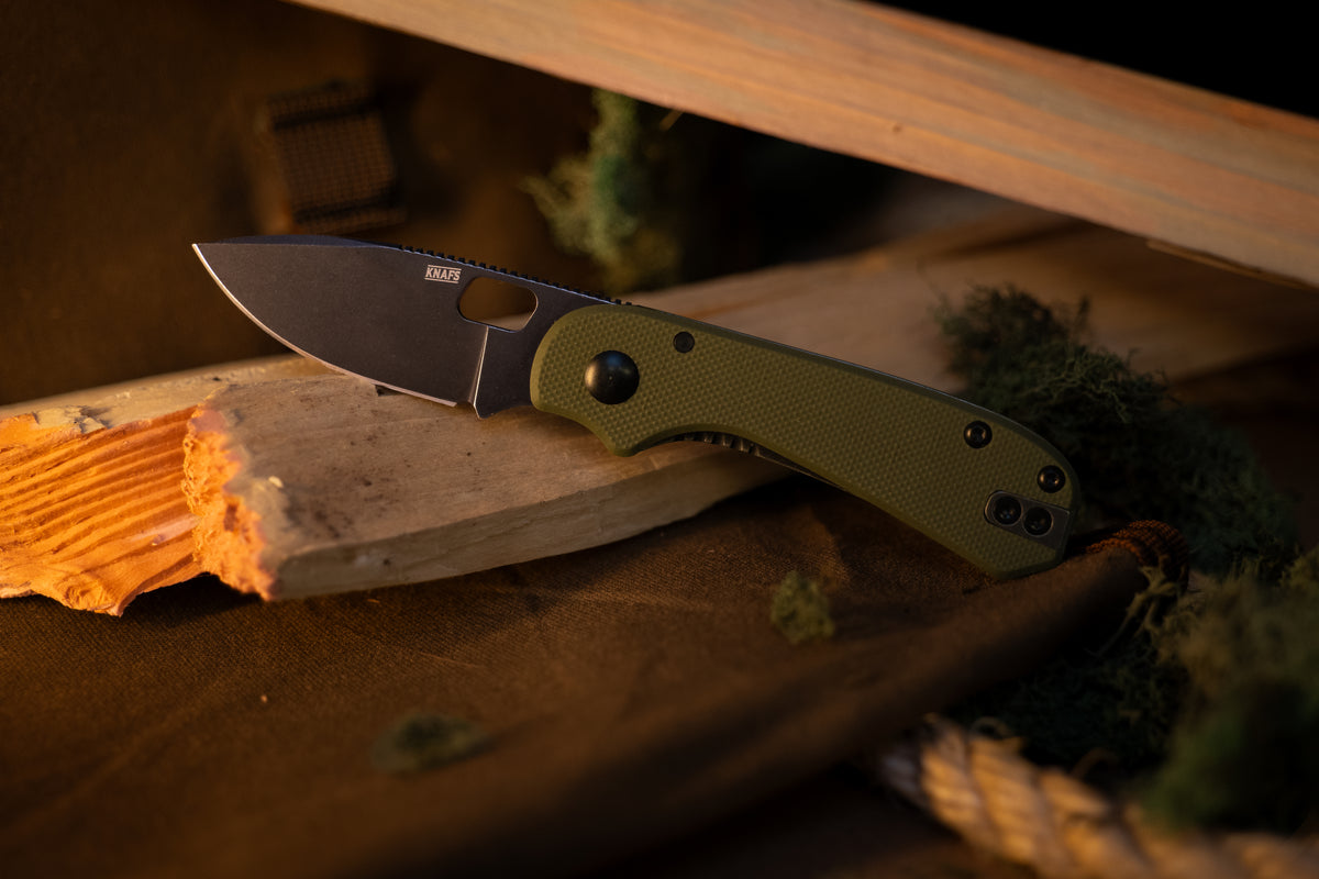 A folding pocket knife with an olive green handle and a dark gray blade, resting on a piece of wood with a rustic background.