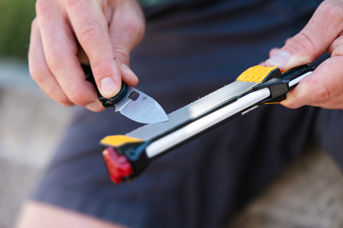 A close-up of a person using a knife to sharpen a metal rod on a black and yellow sharpening stone, with dark shorts and a blurred outdoor background.