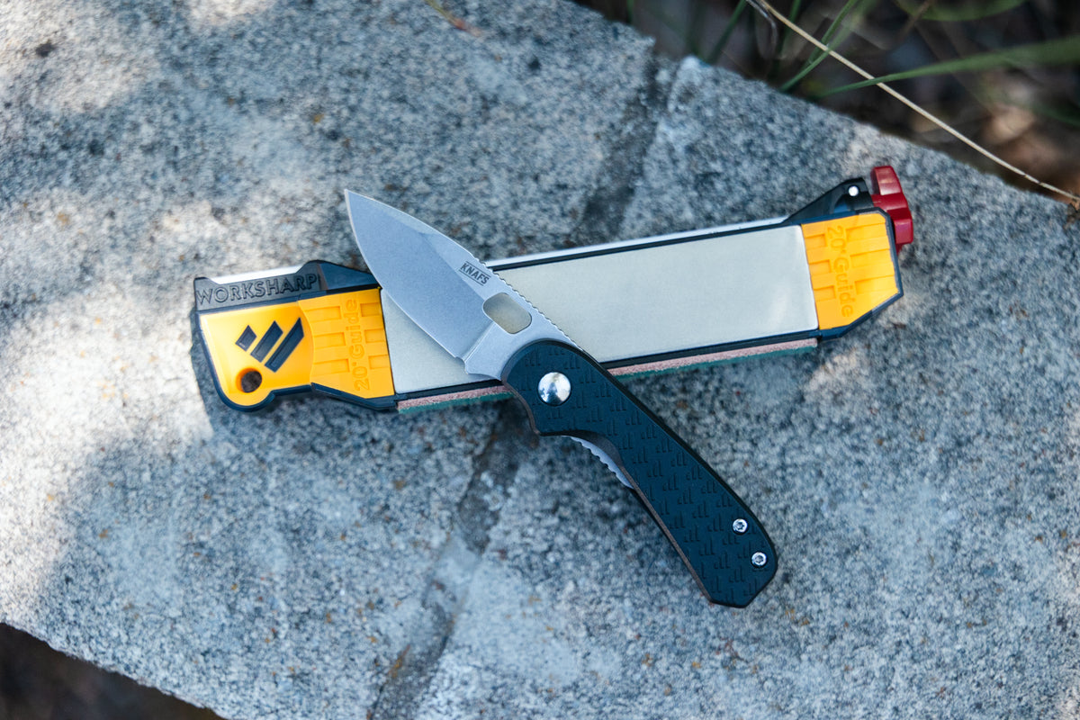 A folding pocket knife with a black textured handle and silver blade lies crossed over a yellow and gray sharpening stone on a gray stone surface outdoors.