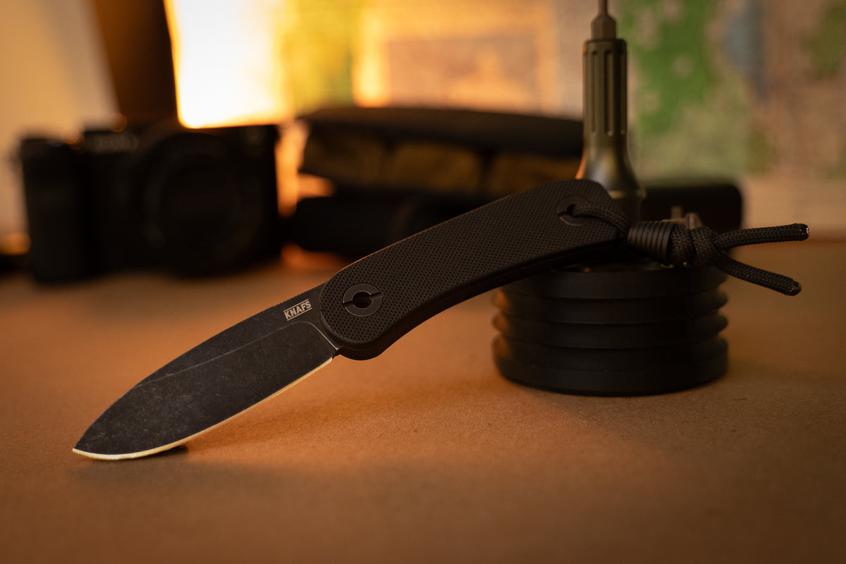 A pocket knife with a textured black handle and a dark gray blade, standing vertically on a surface with a blurred green and warm-toned background.