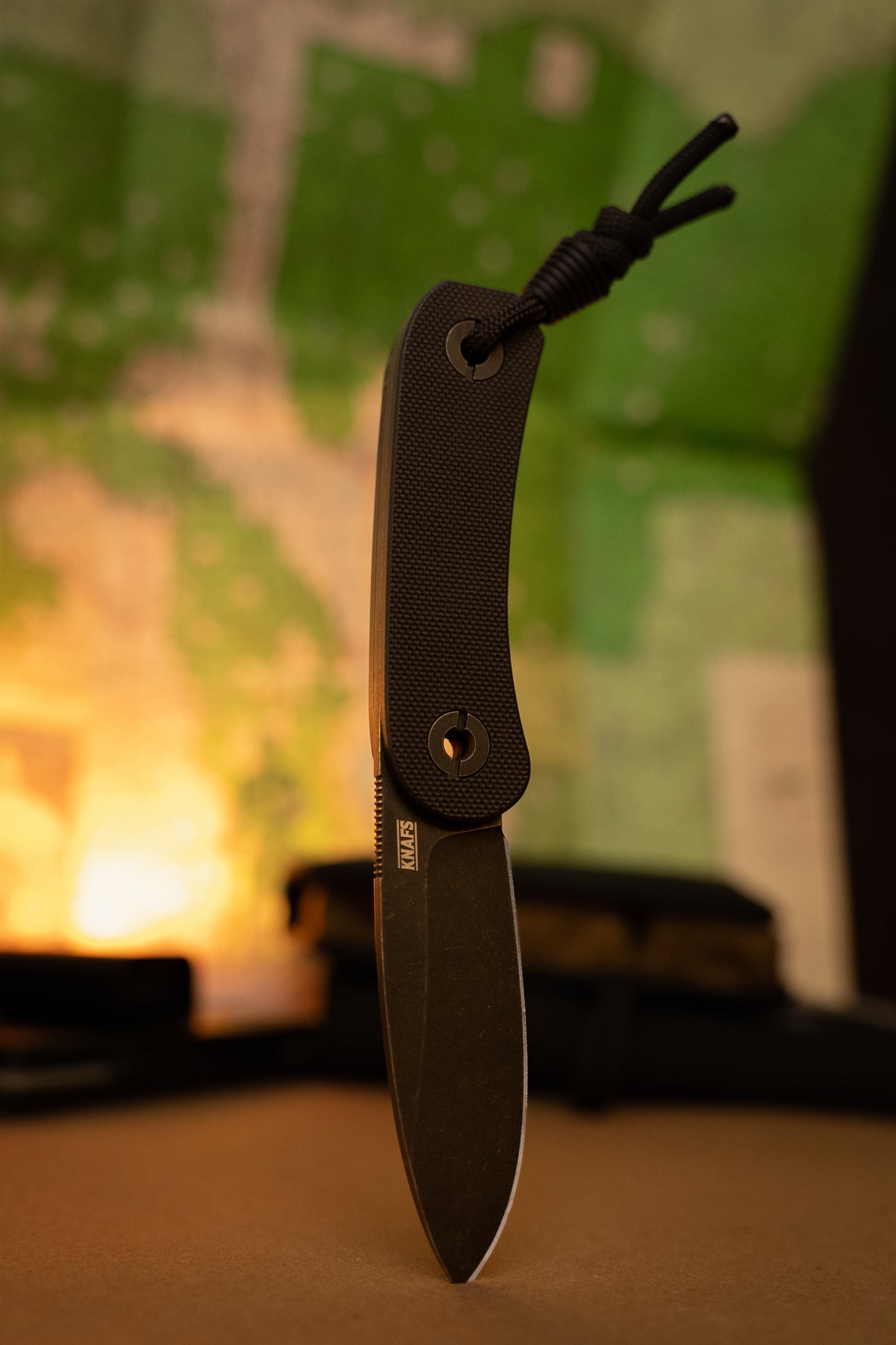 A pocket knife with a textured black handle and a dark gray blade, standing vertically on a surface with a blurred green and warm-toned background.