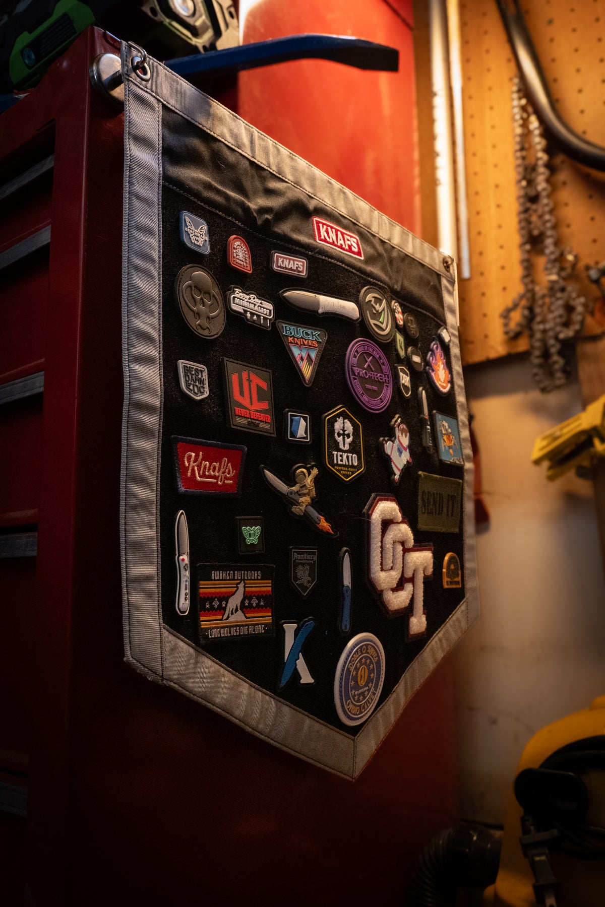 A black canvas wall-mounted patch pocket with gray trim, decorated with numerous automotive and motorcycle-themed embroidered patches, hanging on a red tool cabinet in a workshop.