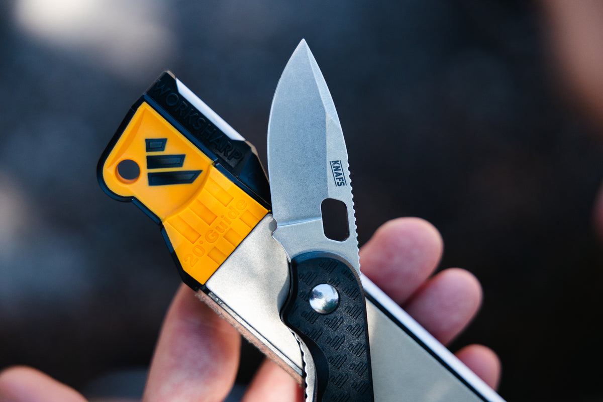 A close-up of a folding pocket knife held in a hand. The knife has a silver stainless steel blade with a black logo, a bright yellow textured handle scale, and a black textured handle grip.