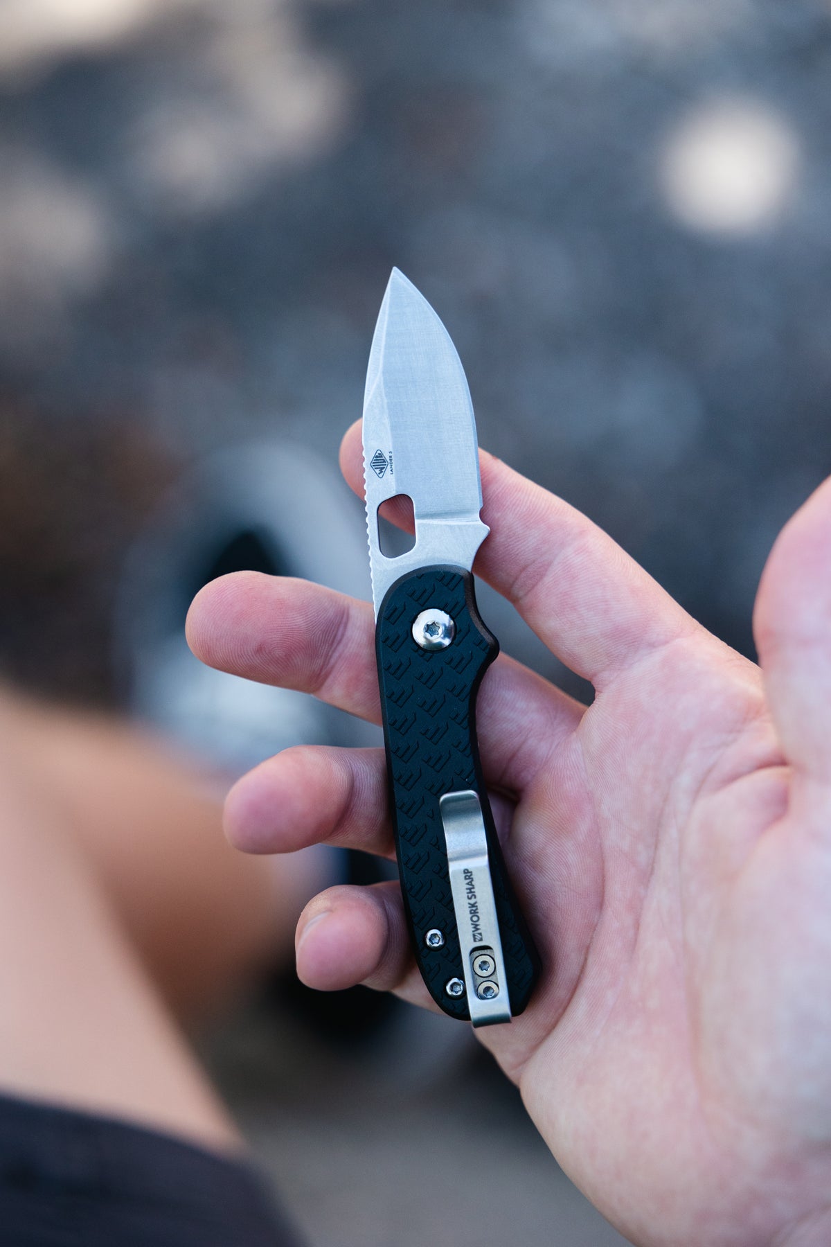 A hand holding a folding pocket knife with a silver blade and a black textured handle featuring a silver clip. The brand 'Worksharp' is visible on the clip.